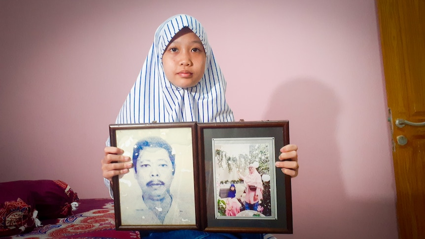 A little girl in a veil holds framed photographs of her late mother and father 