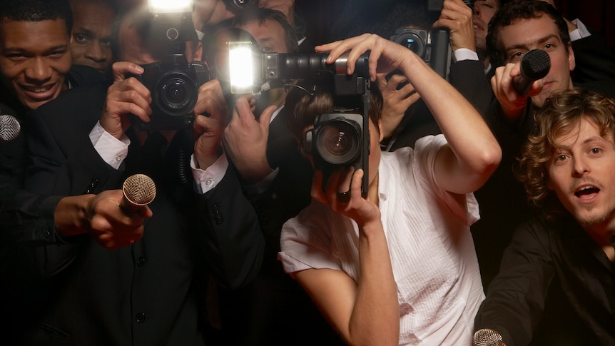 Image of many paparazzi holding cameras pointed at the viewer. Some of the cameras are flashing. 