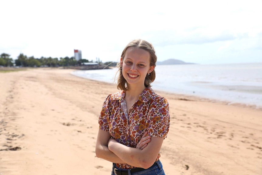 A young woman stands on a beach on a sunny day smiling with her arms crossed.