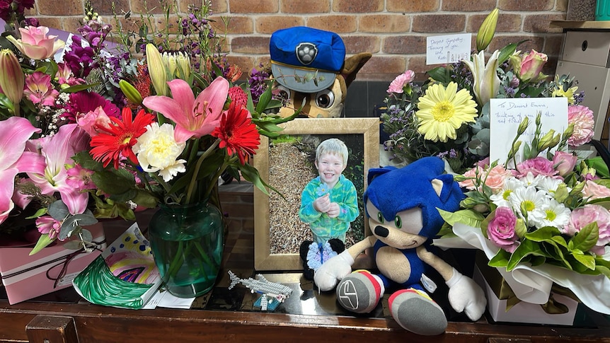 Flower arrangements surround a photograph of a small boy on a table.