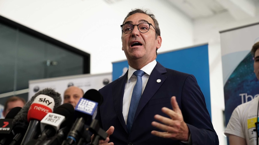 A man wearing a suit speaks to microphones on stands