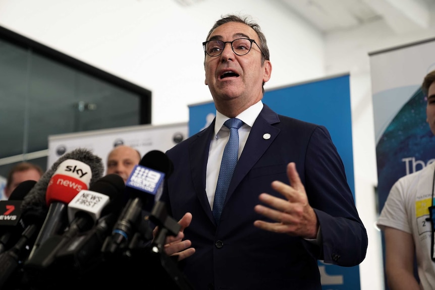 A man wearing a suit speaks to microphones on stands