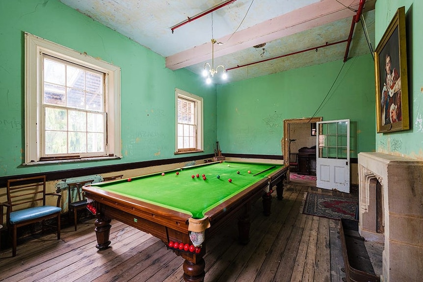 An old room with peeling paint containing a pool table and a fire place.
