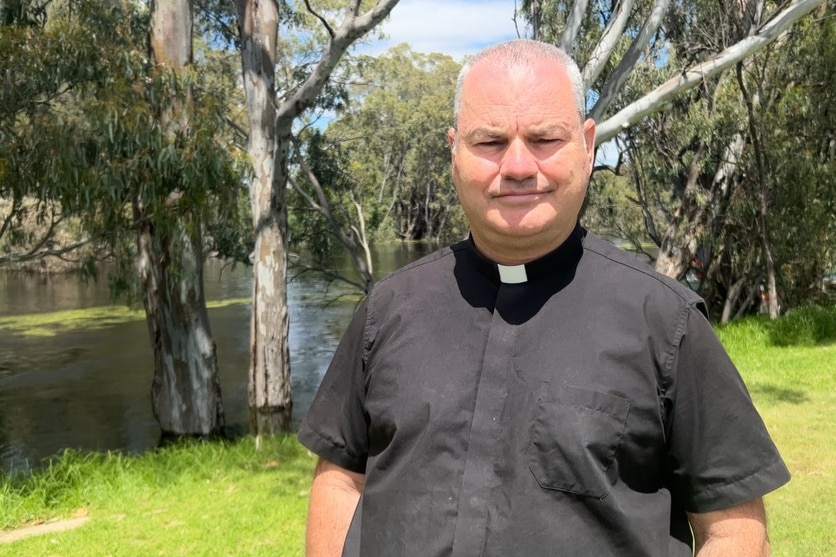 A priest wearing a white collar stands next to a river with trees behind him.