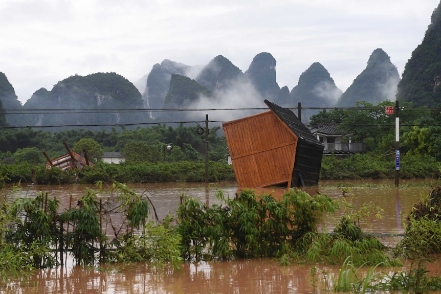 A wooden building sits on its side in floodwater with karst mountains in the background.