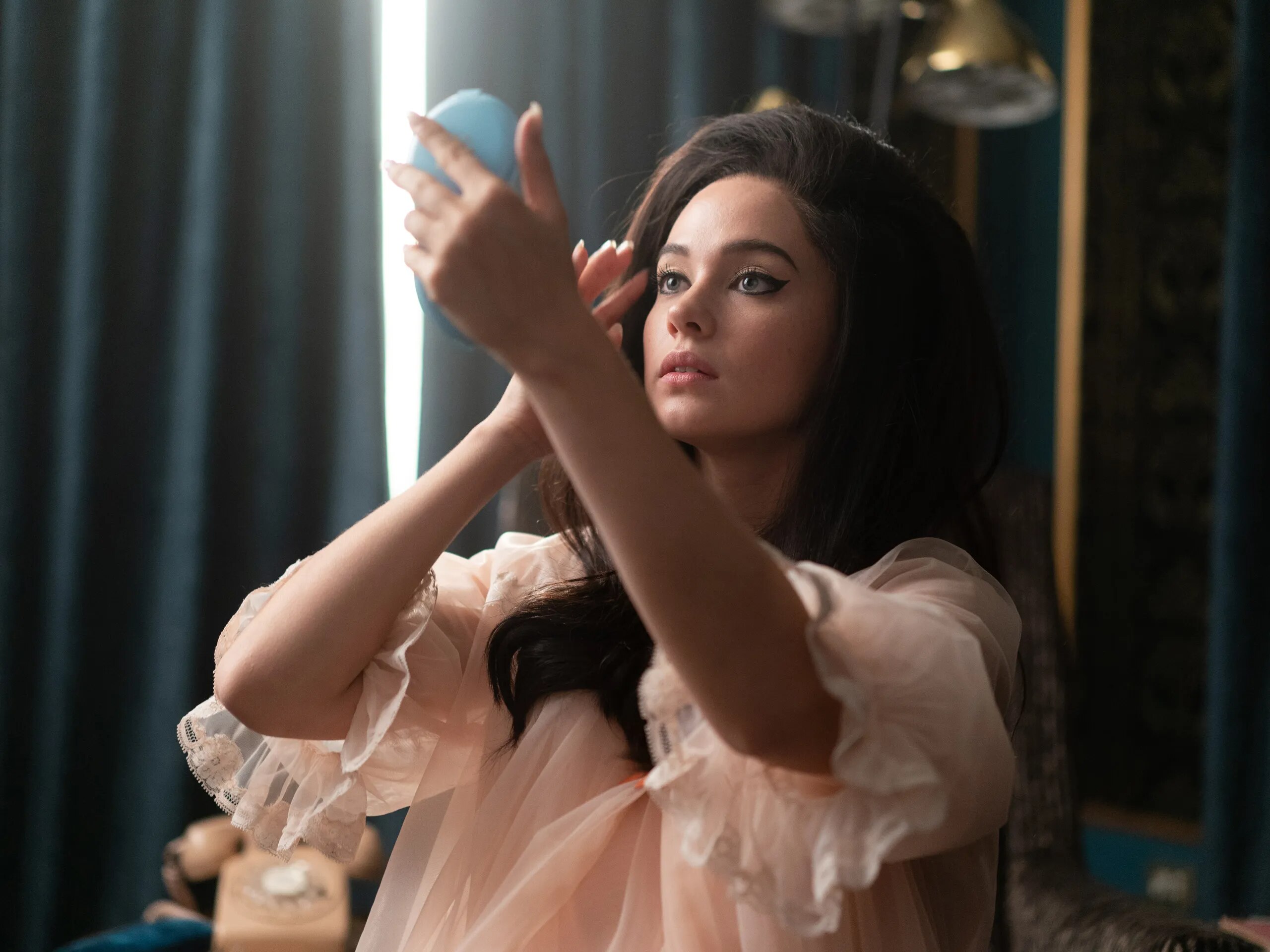 Cailee Spaeny as Priscilla Presley in the film Priscilla. She is wearing a baby pink negligee and looking into a hand mirror