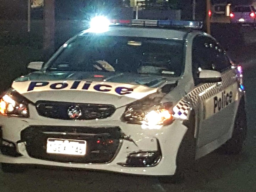 A WA Police car sits on a road with its front smashed up and lights on at night.