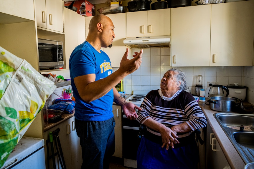 A man stands, gesturing with his arm as he talks to an elderly woman sitting in a kitchen.