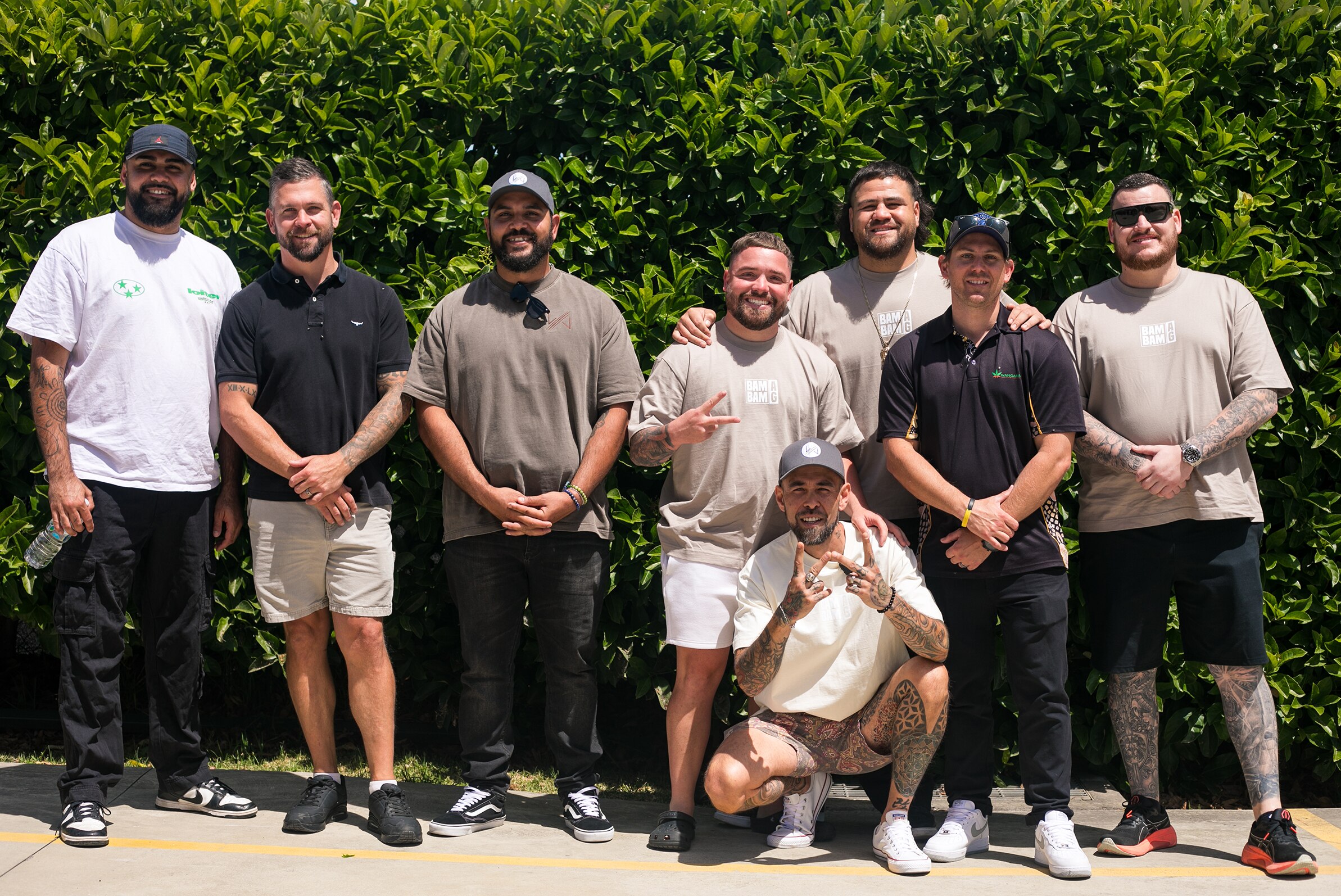A group of men stand outside in front of a hedge - some are wearing caps and one person is kneeling & pose for camera