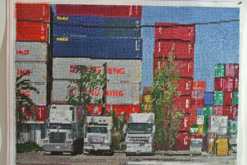 Needlepoint depicting colourful, stacked shipping containers with three large trucks lined up in front of them.