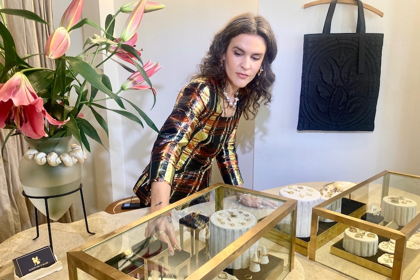 A jewellery store owner in her thirties fixes up a display of gold rings on a display table, there are bright flowers on the de