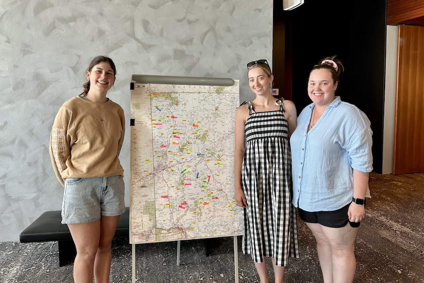 three women smile at the camera. They are standing next to a large map.
