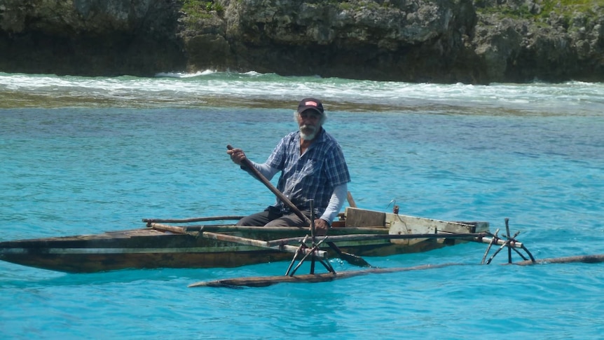 Elderly man paddles in a waka on a bright blue water surrounded by rock. 