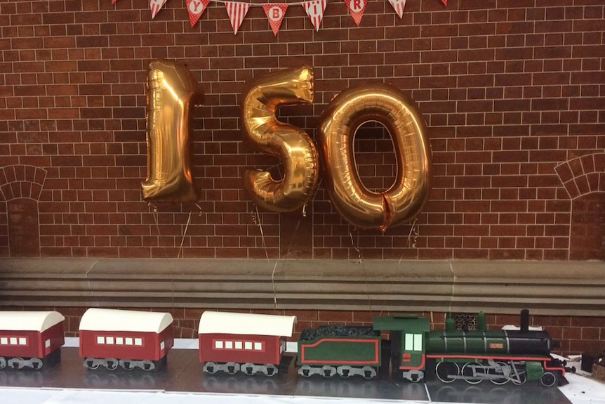 A train cake sits on a table with balloons spelling out 150 above the cake.
