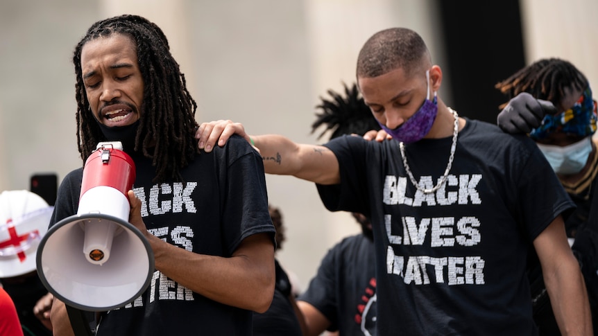 Two sad-looking men wearing Black Lives Matter t-shirts. One has his hand place on the other in prayer.