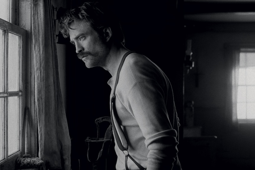 Black & white image from the movie the Lighthouse with Robert Pattinson as a young lighthouse keeper looking out a window