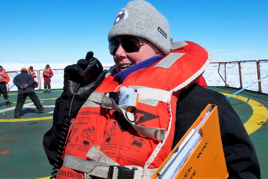 Close up of a woman on ship surrounded by ice, wearing a lifejacket and grey beanie speaking into walkietalky