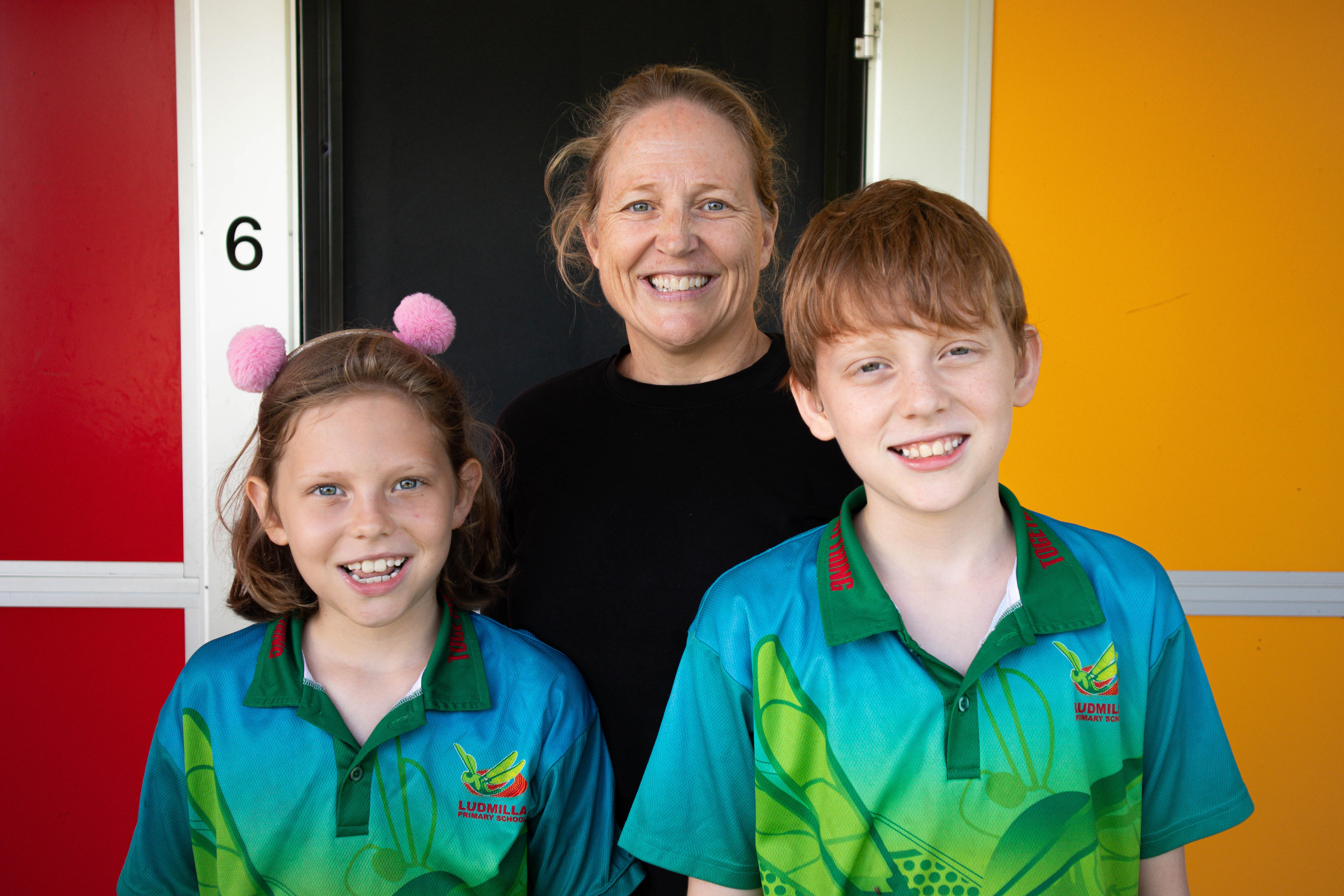 A woman smiles at the camera, in front of her are two happy kids in Ludmilla Primary uniforms.
