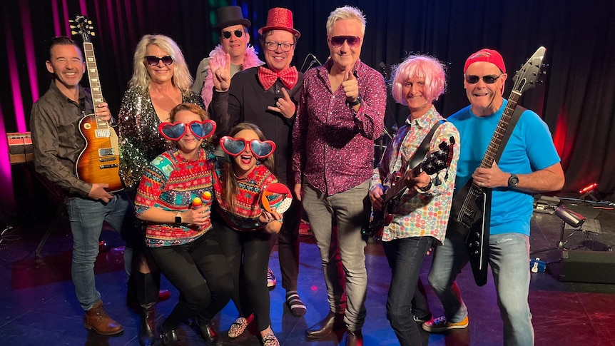 ABC Band photo of nine performers, two holding guitars with two women in christmas jumpers and heart-shaped glasses