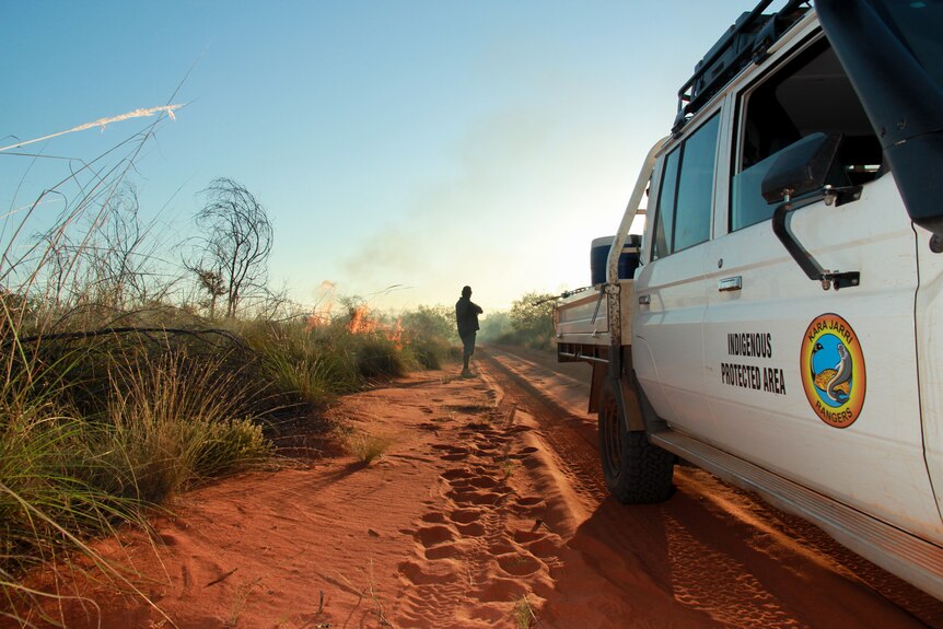 A vehicle on a red sandy track with a man next to a fire.