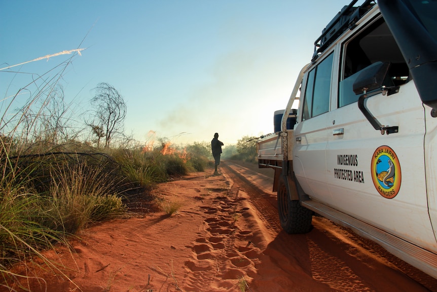 A vehicle on a red sandy track with a man next to a fire.