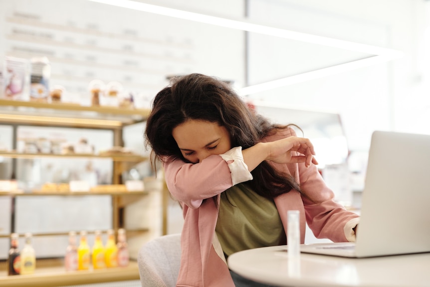 Woman coughing or sneezing into her elbow at her desk with laptop in front of her