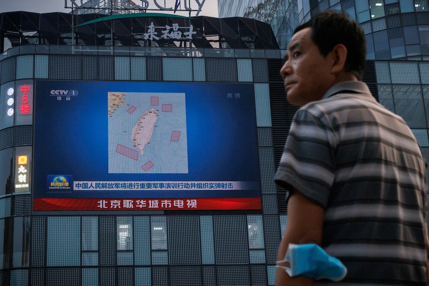 Man stands in front of large screen on building shows Chinese news broadcast with a map of Taiwan.