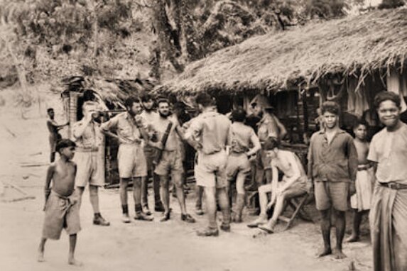 A black and white photo of off-duty soldiers mingling with local Timorese people outside a thatched-roof hut.