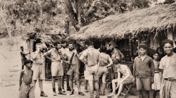 A black and white photo of off-duty soldiers mingling with local Timorese people outside a thatched-roof hut.