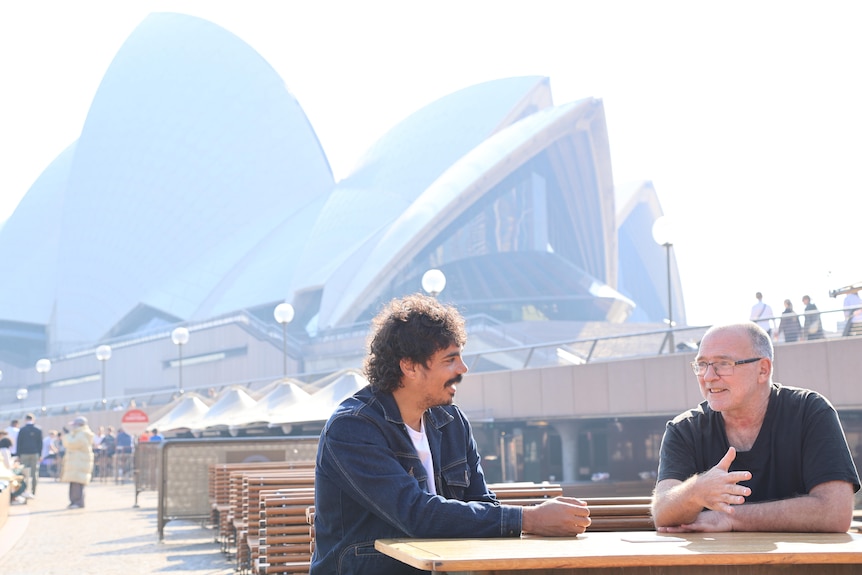 Two men sit at a cafe smiling and talking with the Sydney Opera House filling the background behind them