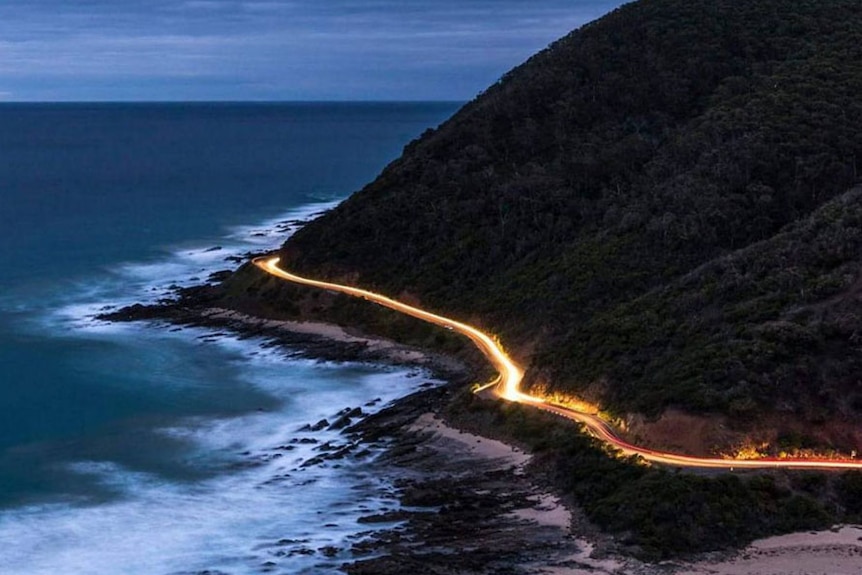 Elevated view of a stretch of the Great Ocean Road at dusk, long exposure turning car headlights into a river of light.