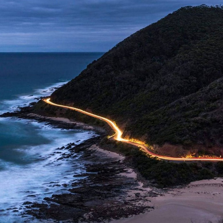 Elevated view of a stretch of the Great Ocean Road at dusk, long exposure turning car headlights into a river of light.