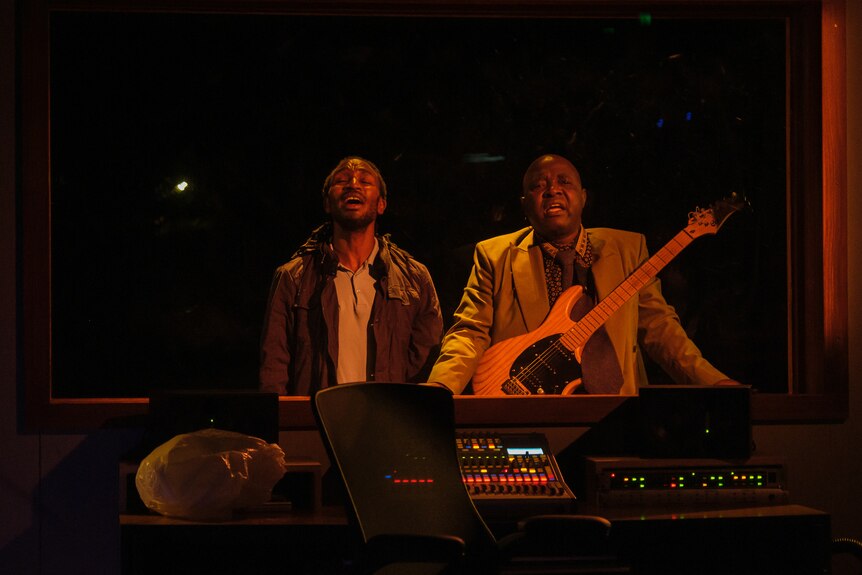 Two African men sing in a soundbooth - one has his dreadlocked hair in a ponytail, while the other is bald and has a guitar