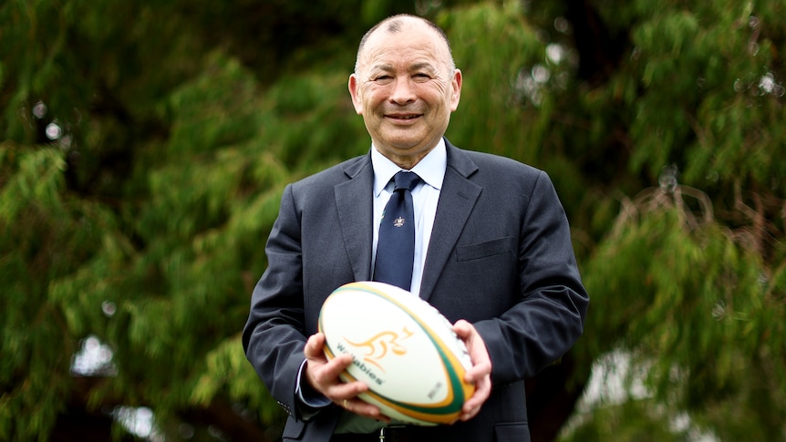 A man in a navy blue suit and tie holds a rugby ball and smiles at the camera with a tree in the background