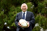 A man in a navy blue suit and tie holds a rugby ball and smiles at the camera with a tree in the background