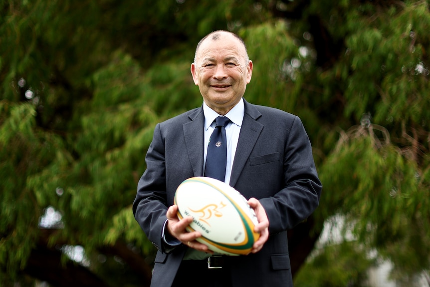 A man in a navy blue suit and tie holds a rugby ball and smiles at the camera with a tree in the background