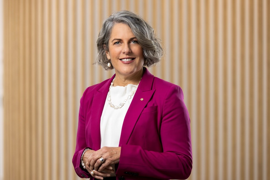 A woman with silver hair, wearing a pink blazer, smiles at the camera