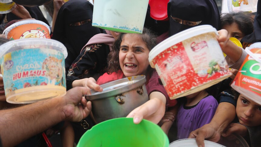 A crowd of women and children reach buckets and bowls towards the camera, a hand in the left corner reaches to give a metal bowl