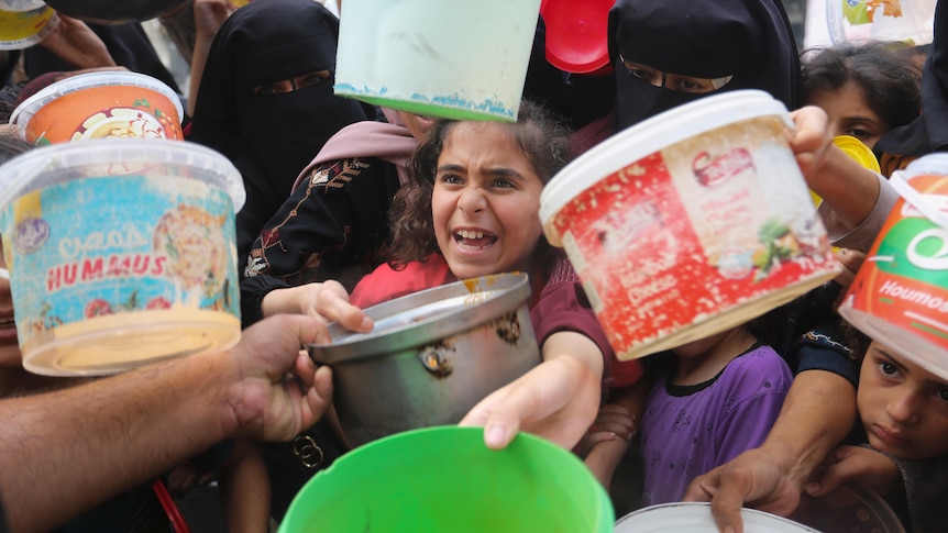 A crowd of women and children reach buckets and bowls towards the camera, a hand in the left corner reaches to give a metal bowl