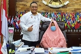 A woman sits in a chair wearing a mask and a pink hijab while a man stands next to her.