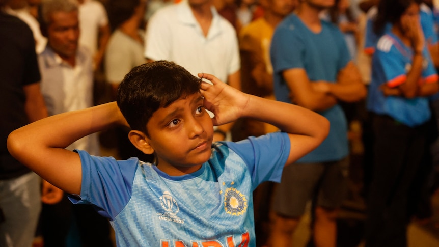 A young Indian boy wearing blue looks sad as he pulls his arms behind him in crowd watching TV