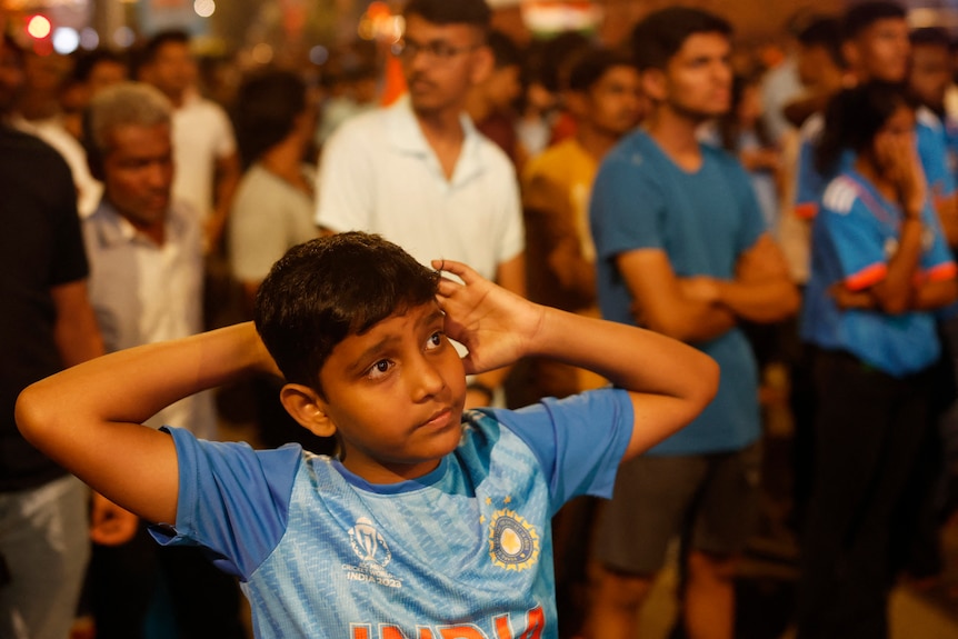 A young Indian boy wearing blue looks sad as he pulls his arms behind him in crowd watching TV
