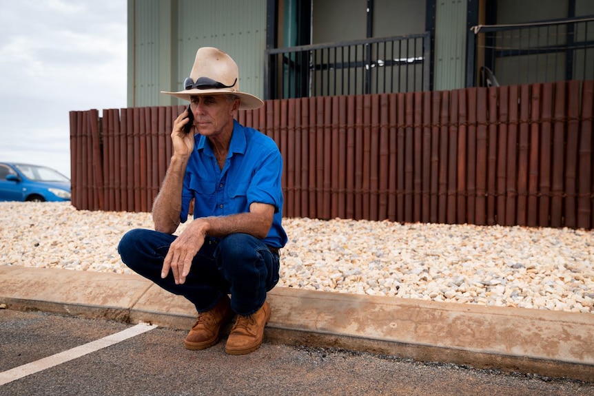 A man in an Akubra hat sits on his heels on a street kerb talking on a phone.