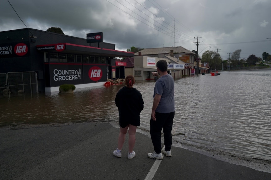 Two people stand looking at rising flood water in Skipton outside businesses. 