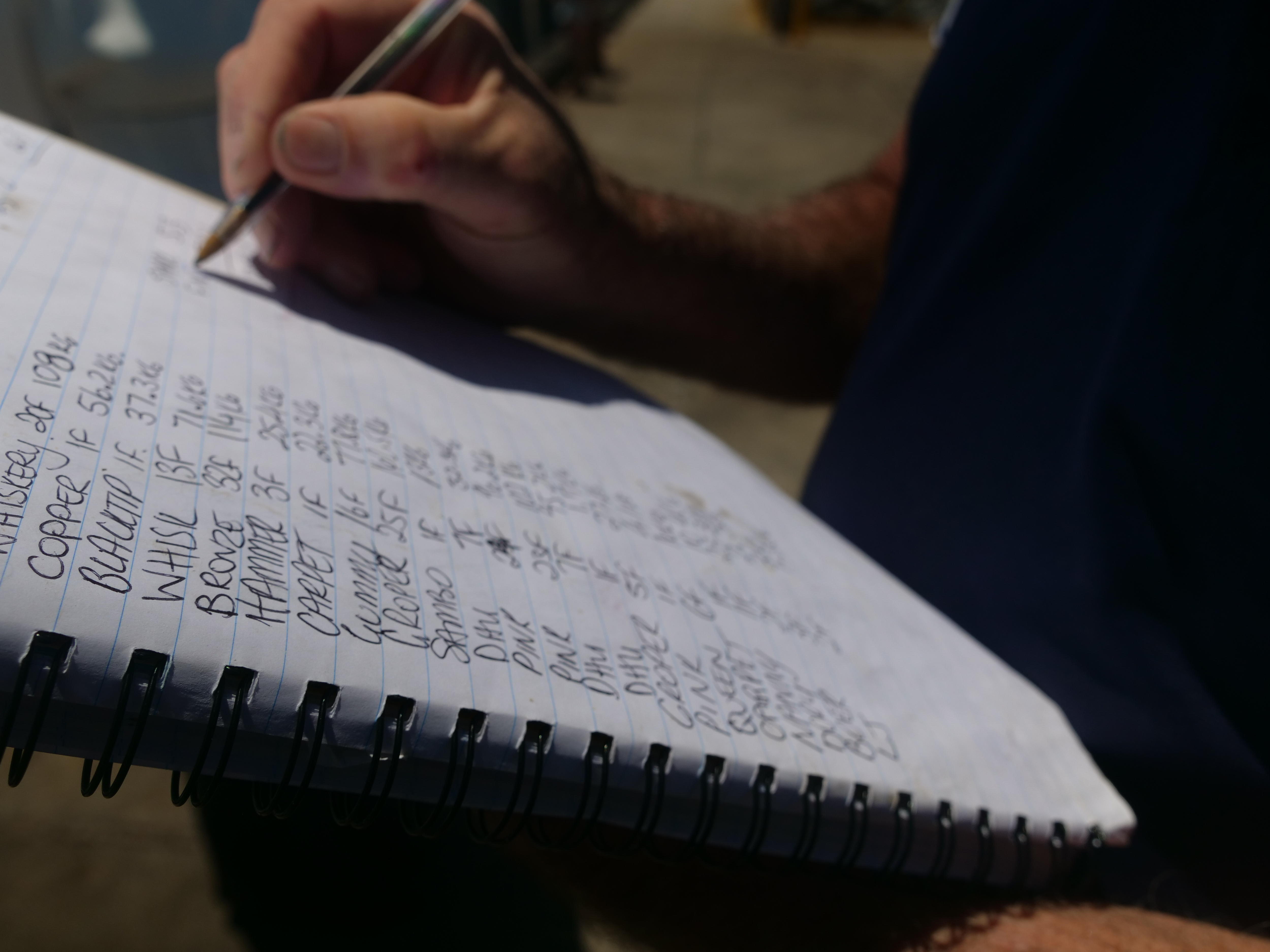 Un primer plano de la mano de un hombre anotando el peso y las especies de peces en un cuaderno rayado con un bolígrafo negro. 