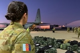 A woman wearing a camouflaged ADF uniform looks over a row of duffel bags and people getting off a plane.