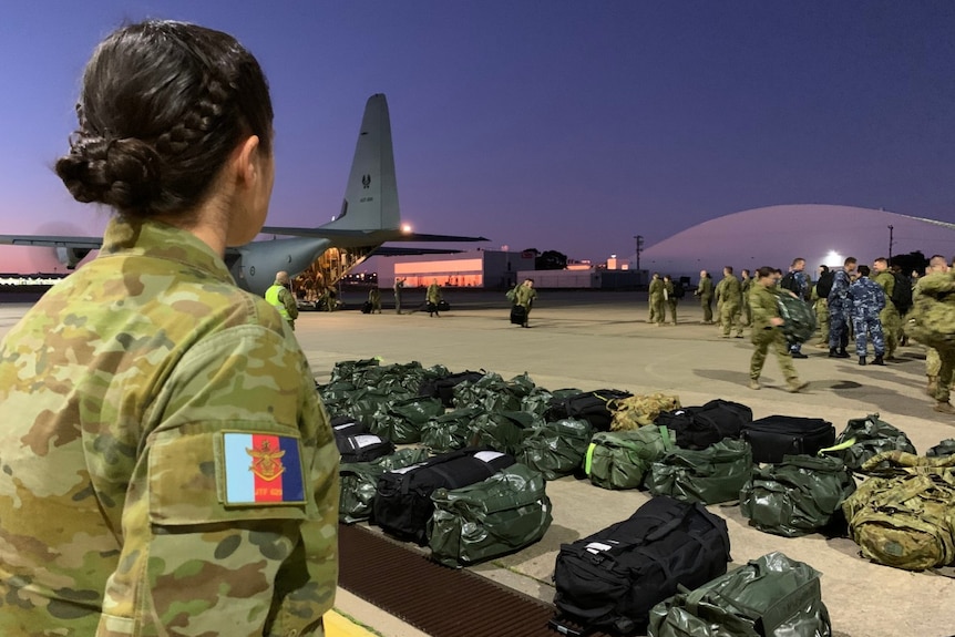 A woman wearing a camouflaged ADF uniform looks over a row of duffel bags and people getting off a plane.