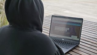A figure wearing a dark hoodie looks at a laptop.