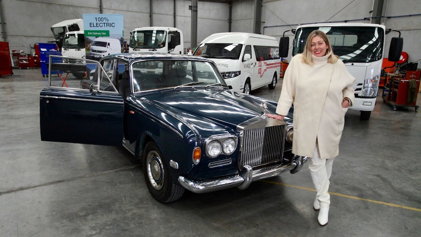 Danielle Wallace stands next to her dark blue vintage 1971 Rolls-Royce in a concrete warehouse.
