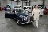 Danielle Wallace stands next to her dark blue vintage 1971 Rolls-Royce in a concrete warehouse.